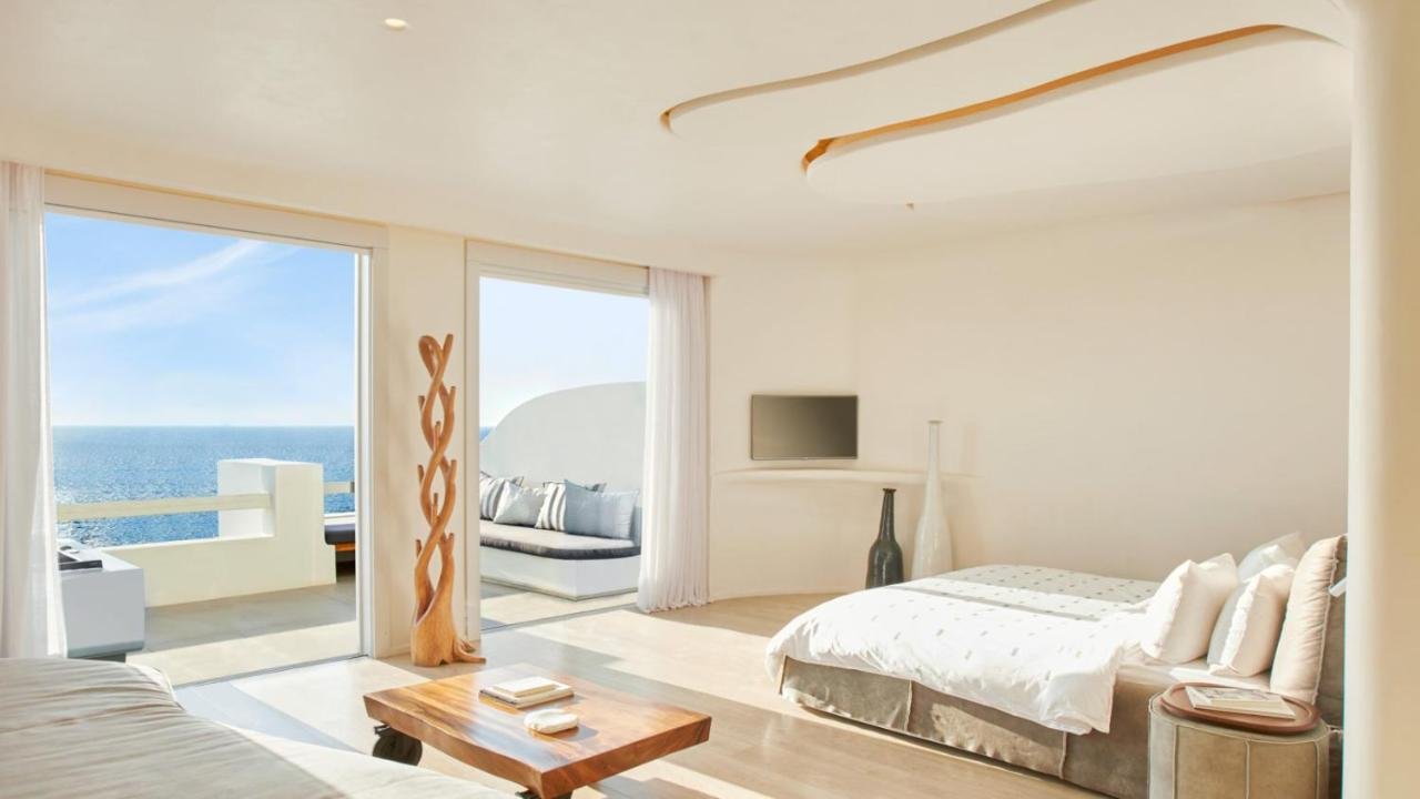 Cavo Tagoo Mykonos suite interior with floor-to-ceiling glass doors open to white terrace and Aegean Sea view
