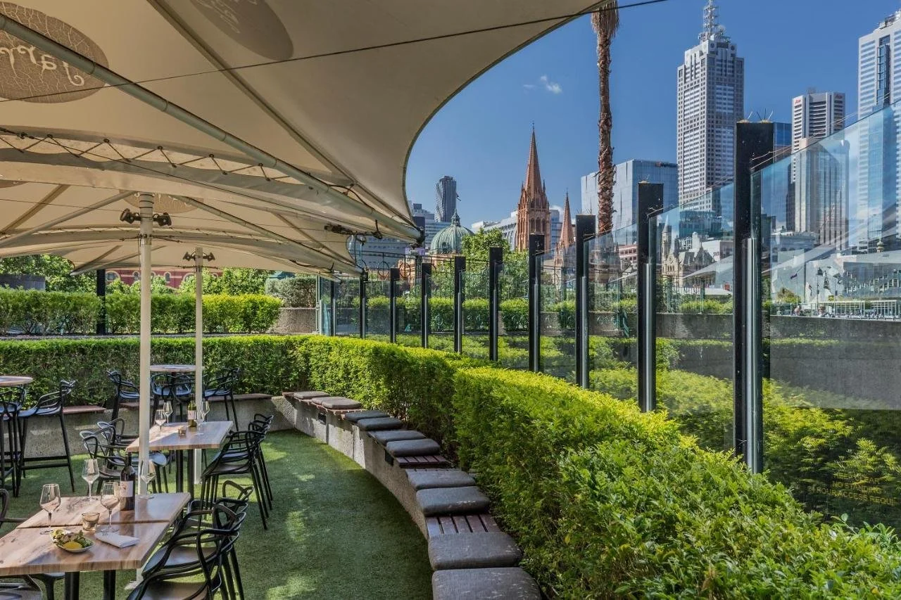 Quay West Suites Melbourne outdoor terrace garden restaurant with St Paul's Cathedral spire, Flinders Street Station dome, and CBD skyline