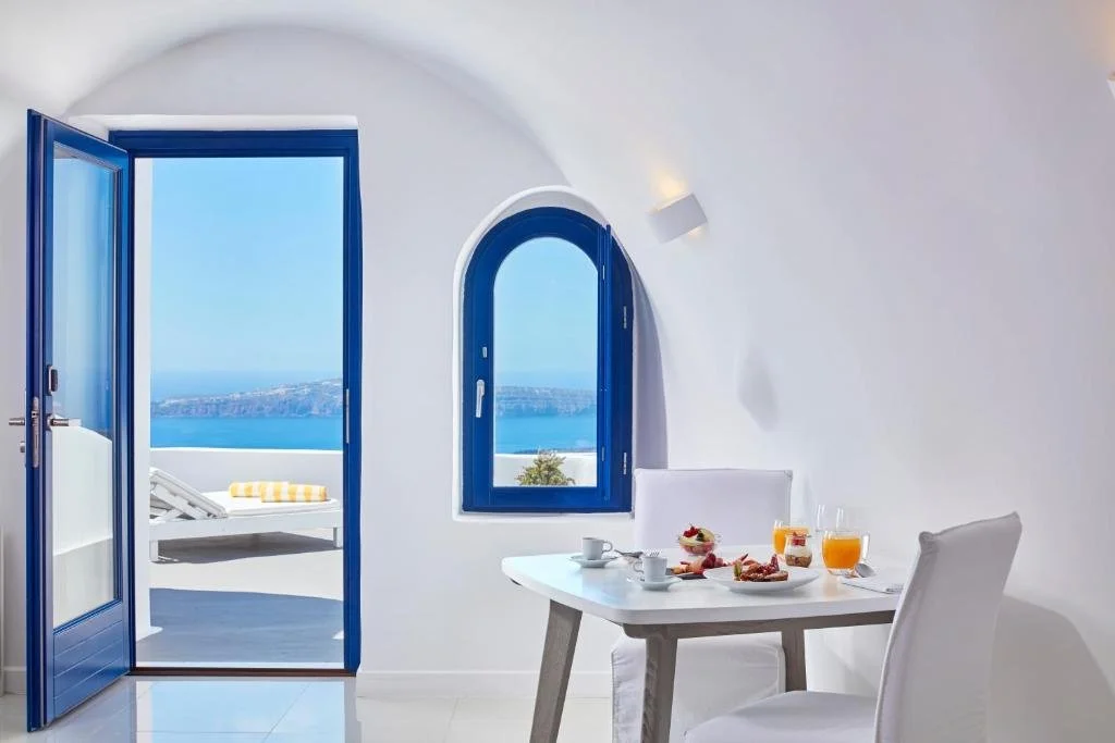 Katikies Chromata Santorini cave-style suite interior with blue-framed door and window open to caldera and volcanic island view