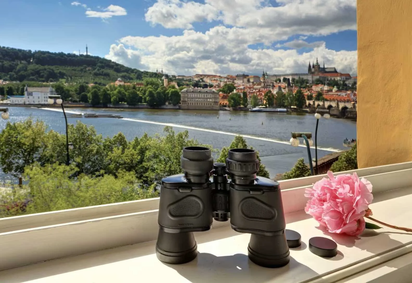 The Mozart Prague binoculars and pink peony on the windowsill with a panoramic view of the Vltava River, Charles Bridge and Prague Castle beyond
