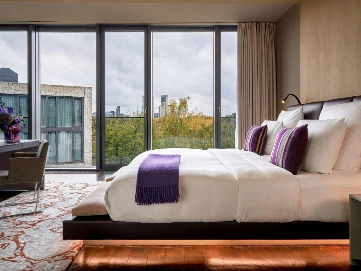 Das Stue Berlin hotel room with platform bed and floor-to-ceiling windows overlooking the Tiergarten autumn tree canopy with the distant Berlin skyline beyond
