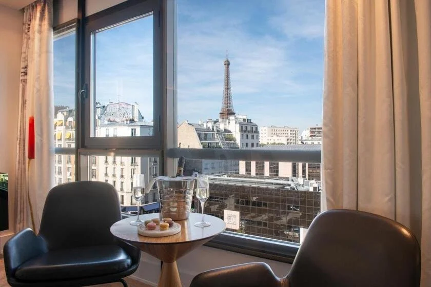 Le Parisis Paris Tour Eiffel hotel room sitting area with two leather chairs, a champagne bucket and macarons on a round table, and the Eiffel Tower filling the window against a blue sky