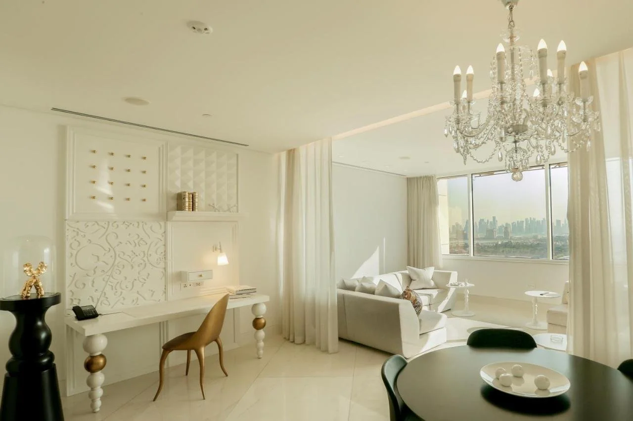 Mondrian Doha Doha all-white suite with crystal chandelier, sculptural wall panels, gold accent chair, and city skyline view through floor-to-ceiling glass