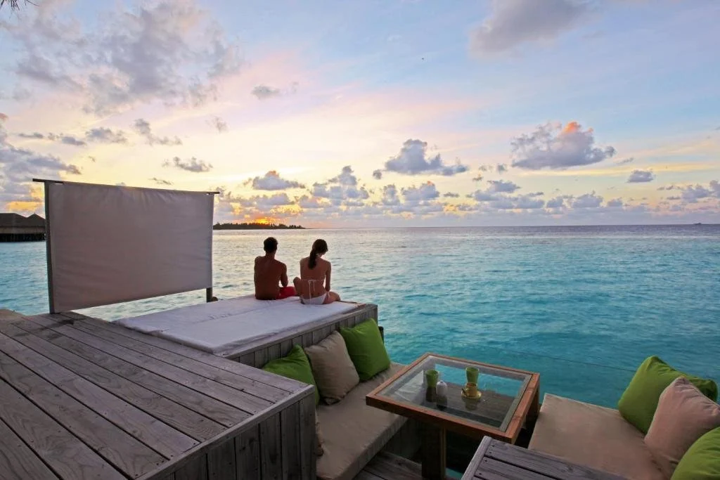 Six Senses Laamu Maldives overwater deck at sunset with two guests seated at ocean edge, green cushion lounge seating and open water horizon