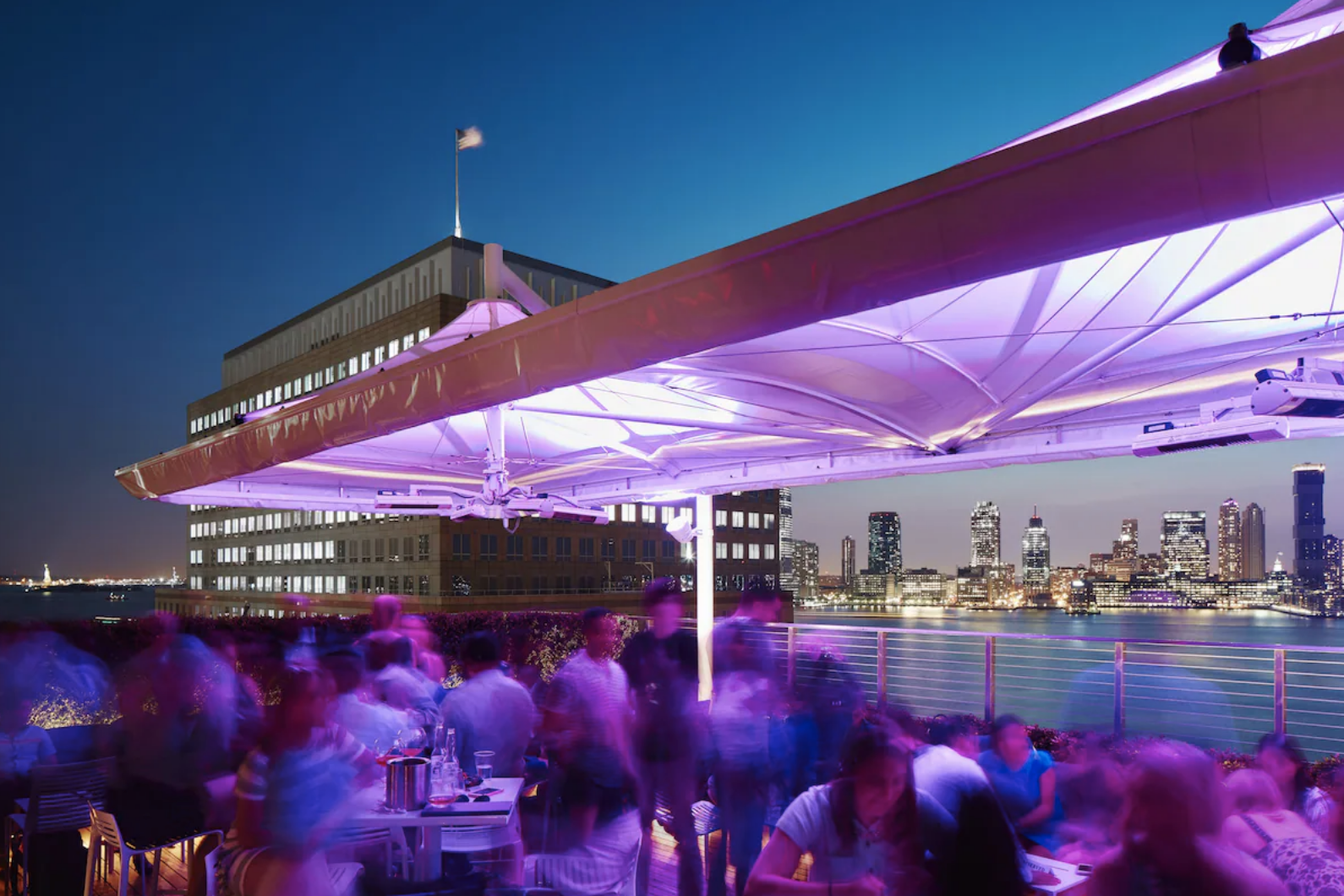 Conrad New York Downtown rooftop bar at night with Jersey City skyline across the Hudson River and Statue of Liberty in the distance