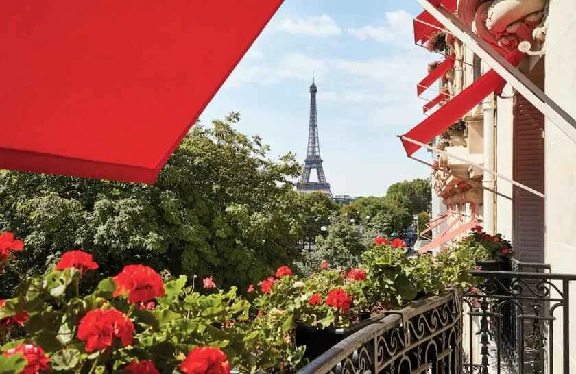 Hôtel Plaza Athénée Paris balcony with red geraniums in flower boxes and a red awning overhead, the Eiffel Tower framed between summer trees in the distance