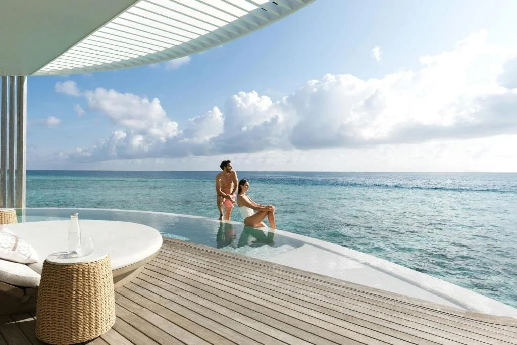Four Seasons Maldives Landaa Giraavaru overwater bar terrace at sunset with woman holding cocktail, illuminated lounge seating and resort lights across lagoon