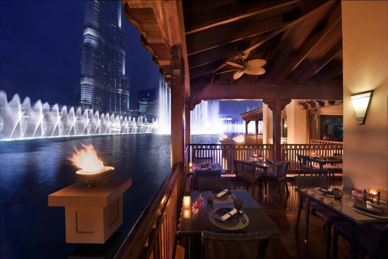 Palace Downtown Dubai Thiptara restaurant terrace at night with fire torch overlooking the Dubai Fountain in full display and the illuminated Burj Khalifa across the lake