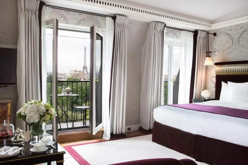 La Réserve Paris hotel suite with a large bed, purple accents, and damask wallpaper, open French doors to a small bistro balcony with the Eiffel Tower visible through the trees in the distance