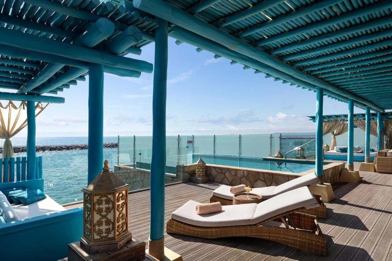 Banana Island Resort Doha by Anantara Doha overwater pool terrace with blue painted pergola, Moroccan lanterns, wicker sun loungers, and an infinity pool merging with the open Arabian Gulf