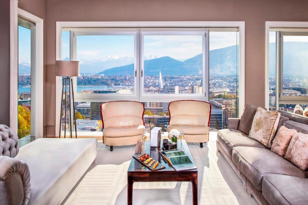 InterContinental Genève Geneva high-floor suite living room with pale upholstered sofas and wraparound windows showing a full bird's-eye panorama of the city, Jet d'Eau, and Alps