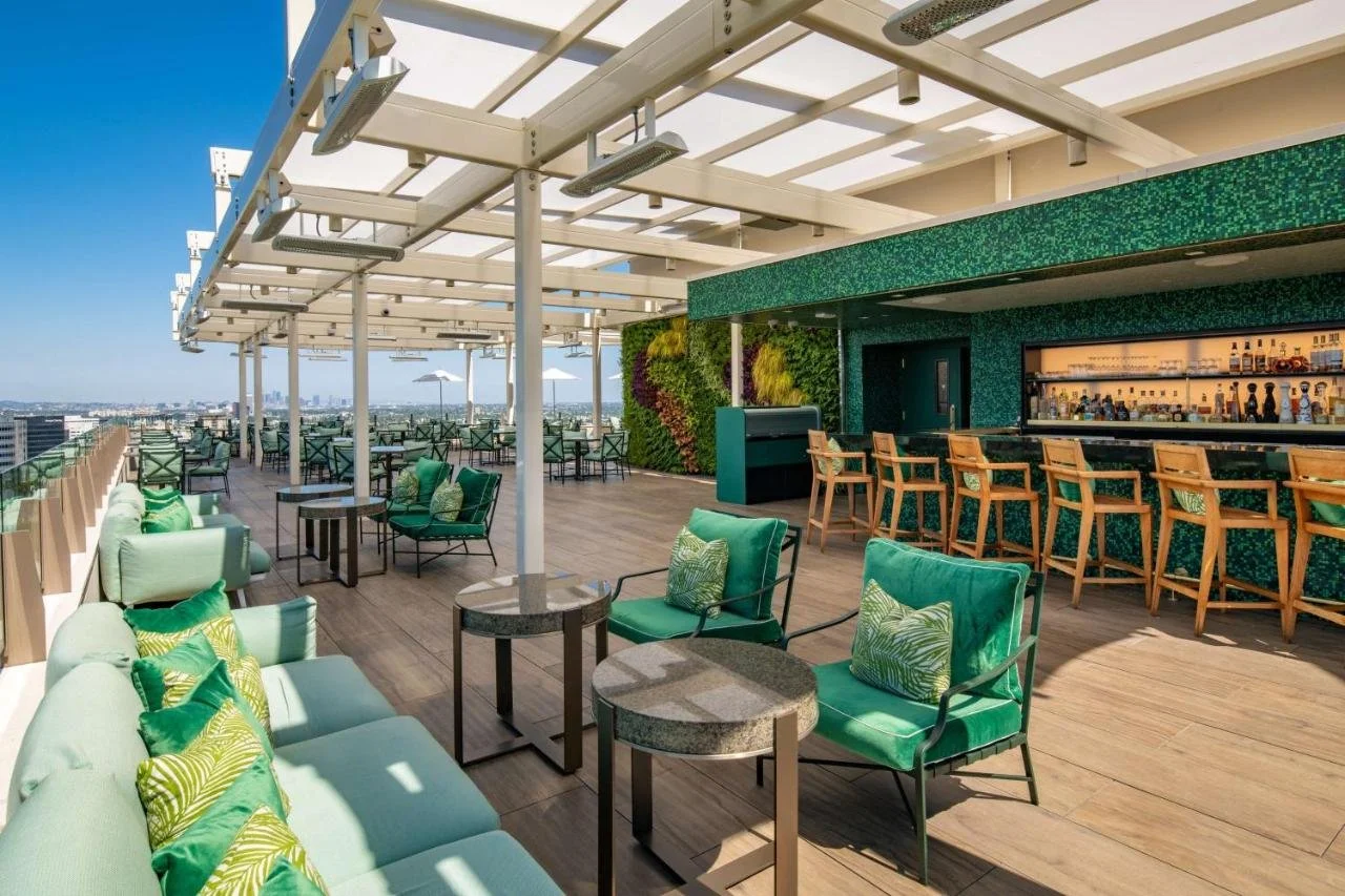 Waldorf Astoria Beverly Hills rooftop bar with green velvet seating, a living plant wall behind the bar counter, and the Los Angeles cityscape stretching to the horizon