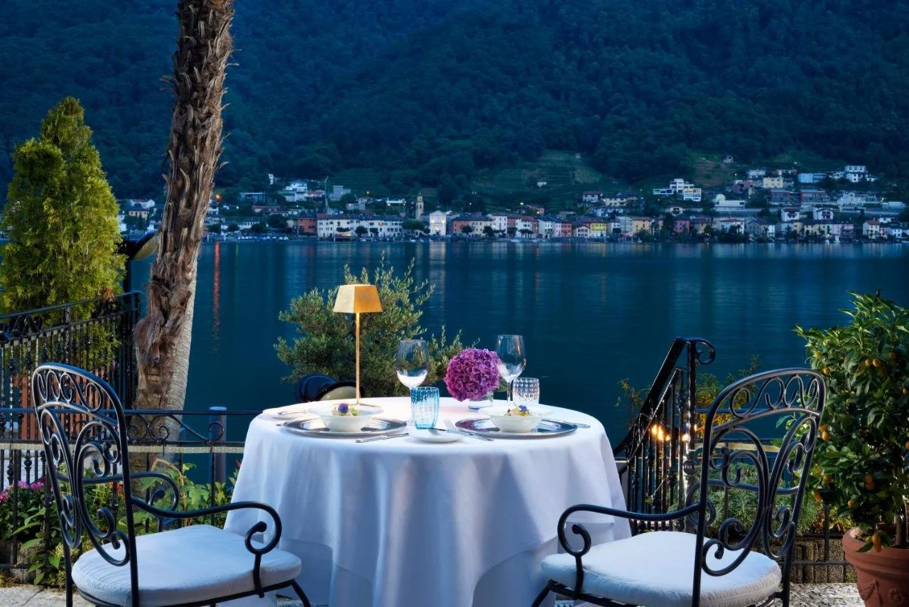 Swiss Diamond Hotel & Spa Morcote evening lakeside terrace with a set dinner table and an illuminated village reflected across Lake Lugano