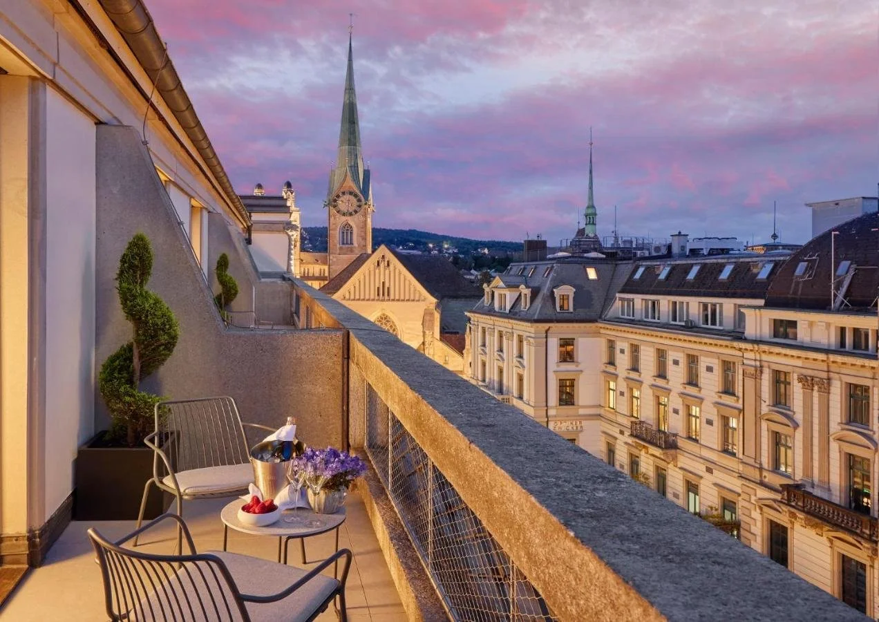 Mandarin Oriental Savoy Zurich rooftop terrace at sunset with champagne and flowers, Fraumünster green spire and Old Town rooftops under a pink sky