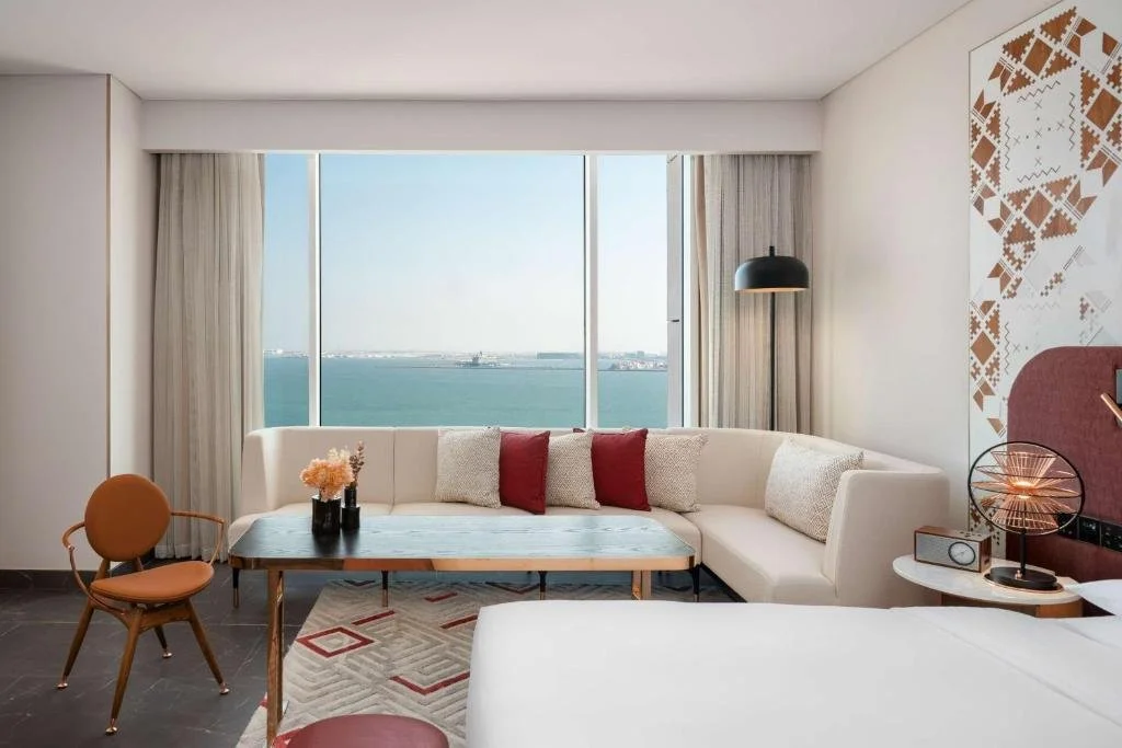 Andaz Doha by Hyatt Doha sea-view room with Al Sadu-inspired geometric wall panel, cream sofa with red cushions, and floor-to-ceiling window overlooking the turquoise Arabian Gulf