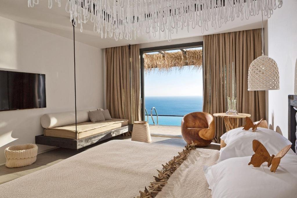 Myconian Utopia Mykonos suite interior with hanging wooden daybed, glass-drop chandelier, and thatched terrace with private pool and Aegean Sea view