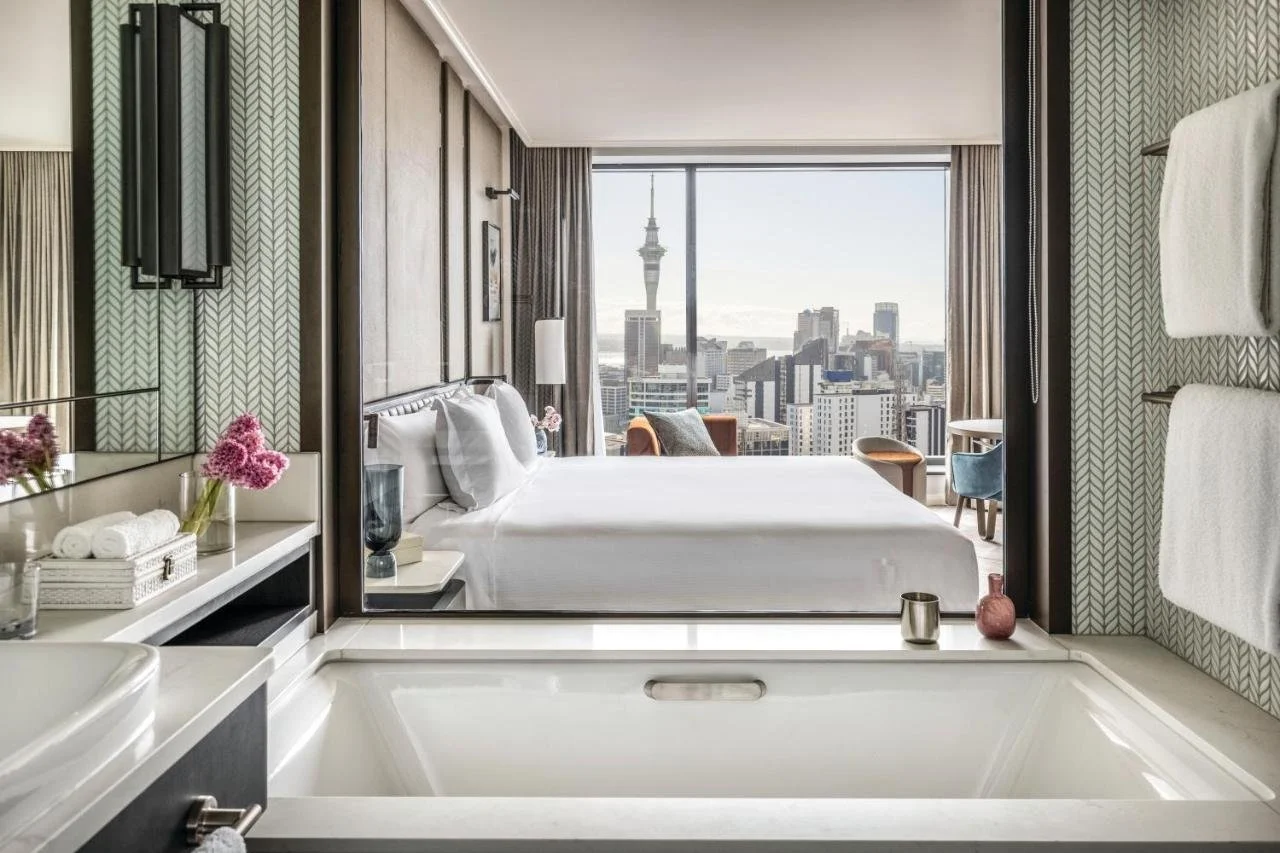 Cordis Auckland Auckland suite bedroom with herringbone-patterned walls and floor-to-ceiling windows on two sides showing the Sky Tower and city skyline in daylight