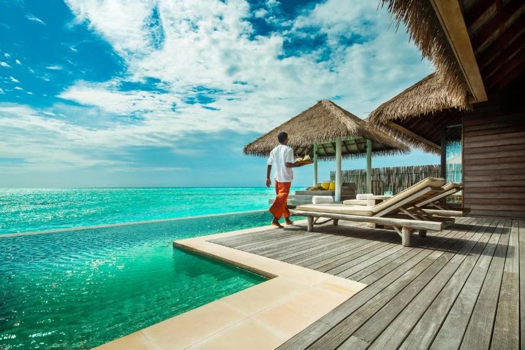 COMO Maalifushi Maldives overwater villa teak deck with private plunge pool, thatched gazebo and staff member carrying tray over turquoise Indian Ocean