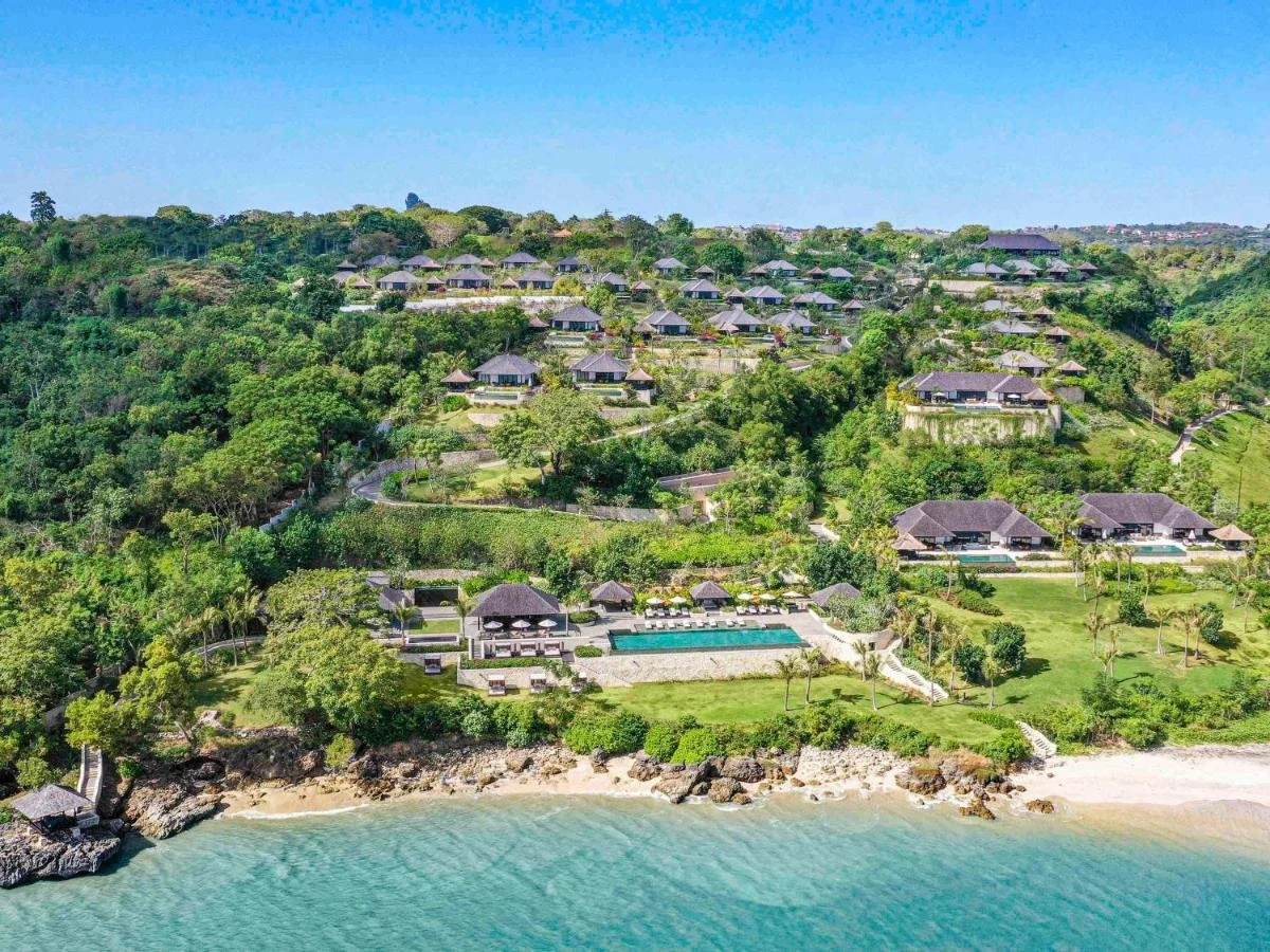 Raffles Bali Jimbaran aerial view of thatched villa resort on hillside with beachfront infinity pool and private beach on the ocean shoreline