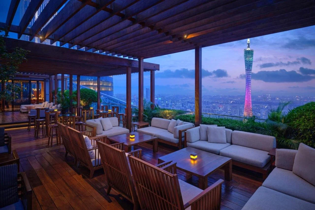Park Hyatt Guangzhou open-air rooftop terrace bar at dusk, wooden pergola and tropical greenery framing the Canton Tower lit in pink and purple against the evening sky
