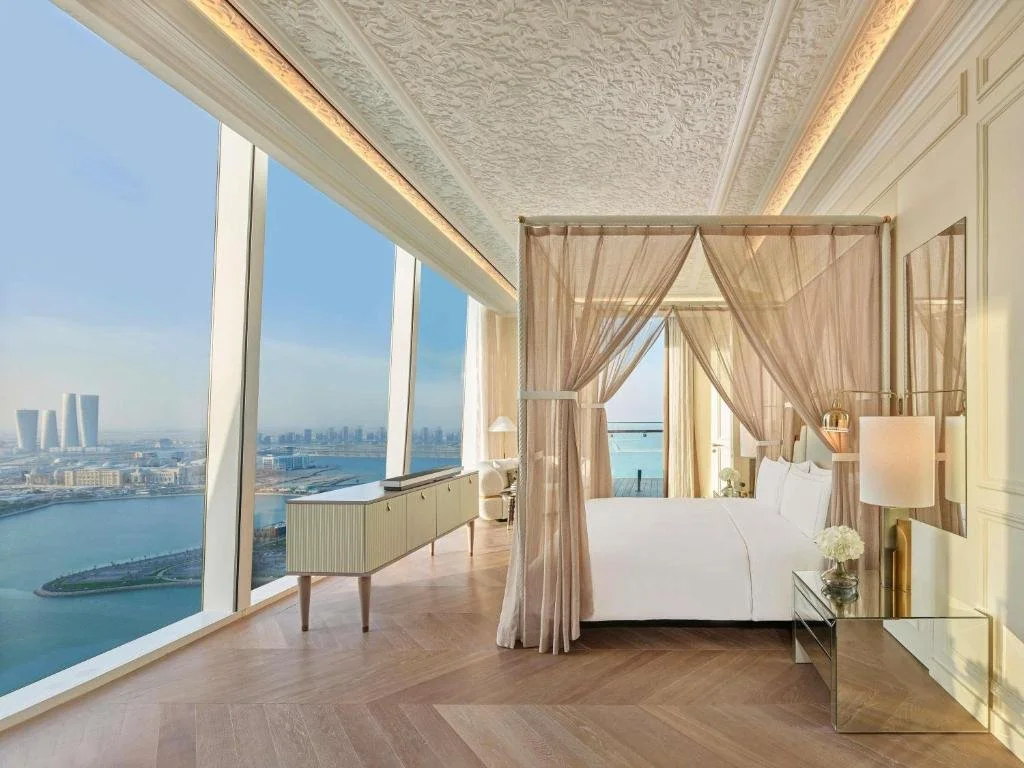 Raffles Doha Doha opulent suite with carved plaster ceiling, gauze canopy bed, herringbone parquet floor, and floor-to-ceiling windows overlooking the turquoise Arabian Gulf and Lusail waterfront