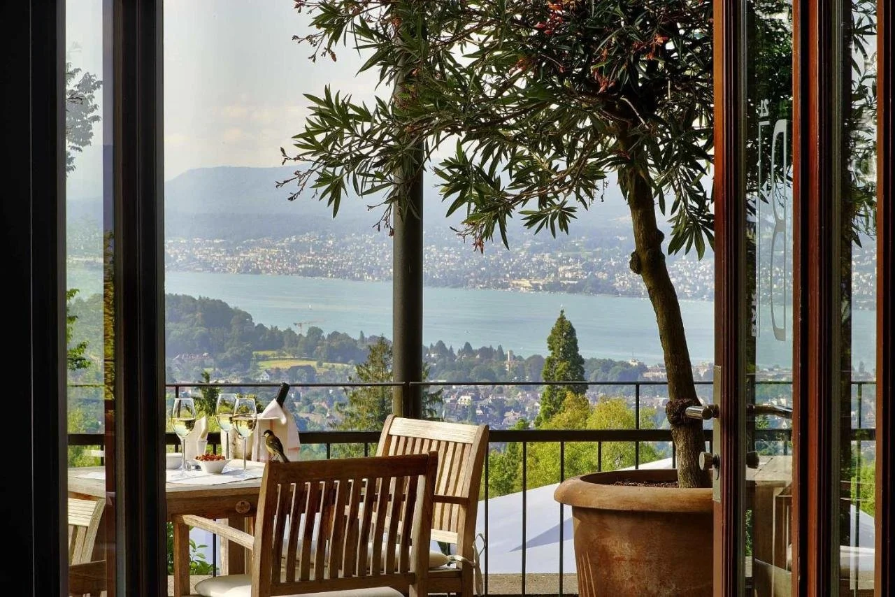 Sorell Hotel Zürichberg Zurich balcony with set table and white wine glasses framed by glass doors, panoramic view of Lake Zurich and forested hills beyond