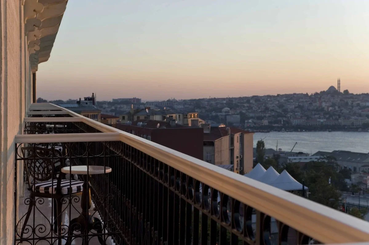 Pera Palace Hotel Istanbul room balcony at sunset with wrought iron railing, Golden Horn waterway and Süleymaniye Mosque silhouette visible on the far horizon