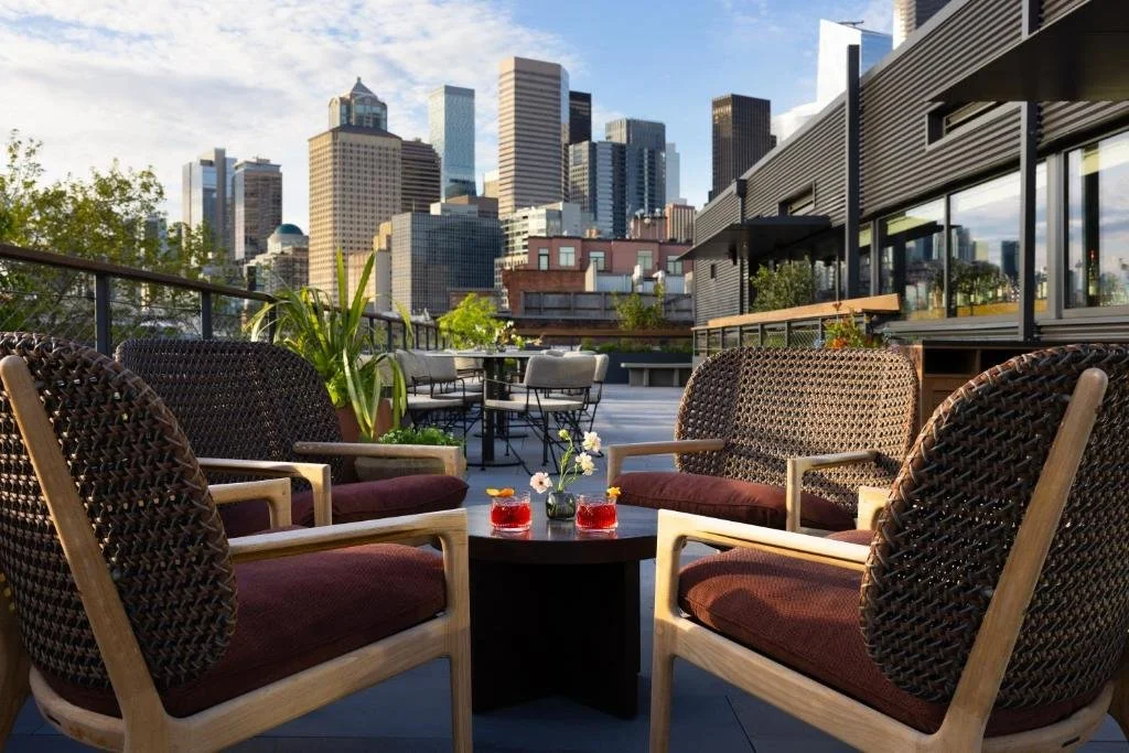 Populus Seattle Seattle Firn rooftop bar terrace with wicker chairs, cocktails on a low table, and an open view of the downtown Seattle skyline under a partly cloudy afternoon sky