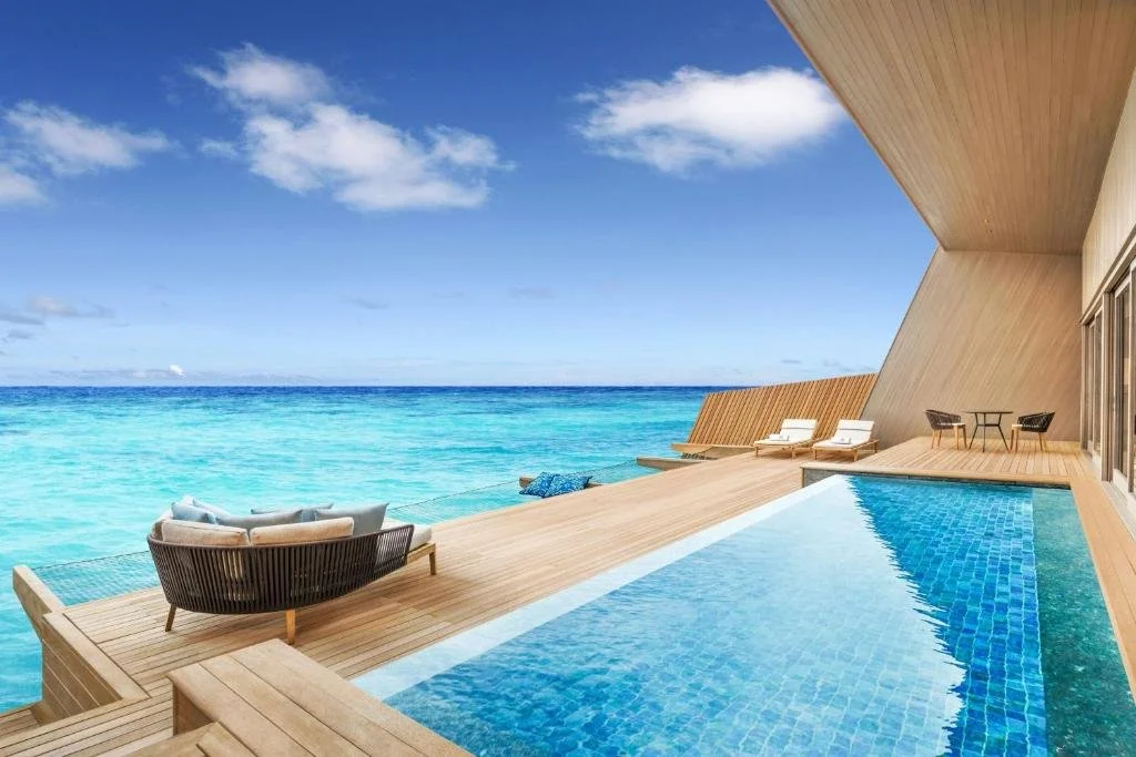 St. Regis Maldives Vommuli Resort overwater villa terrace with long lap pool, timber deck chairs and angular contemporary architecture over turquoise ocean