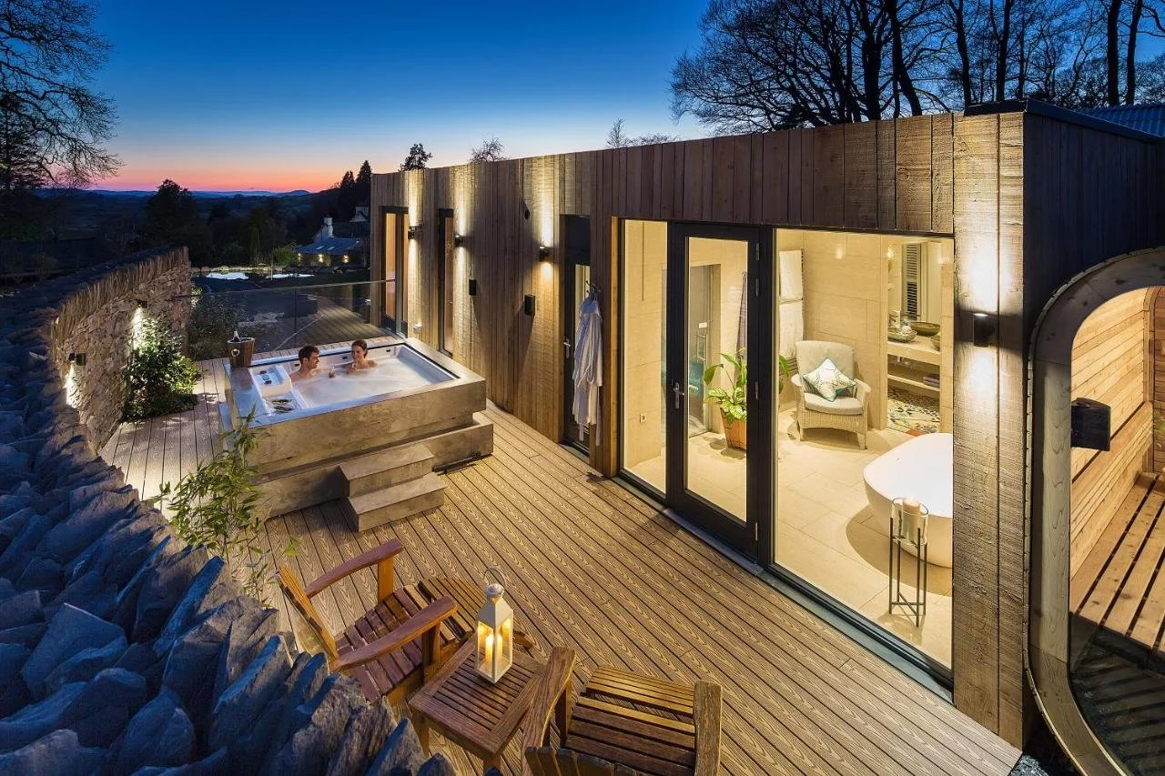 Gilpin Hotel Lake District spa lodge outdoor deck at dusk with stone-built hot tub, wooden terrace, lit suite interior and barrel sauna, Lakeland hills at sunset