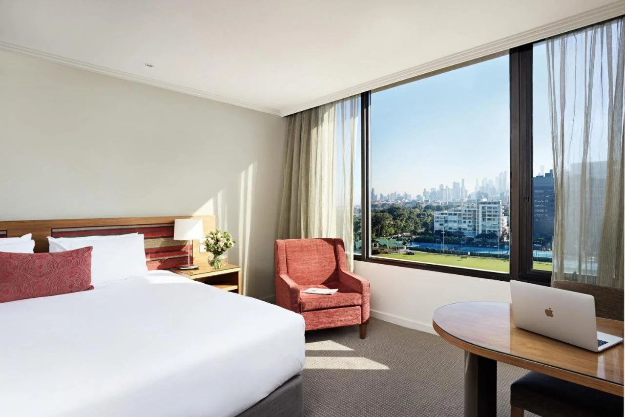 View Melbourne hotel room with large window showing Albert Park sports courts in the foreground and the Melbourne CBD skyline in the distance