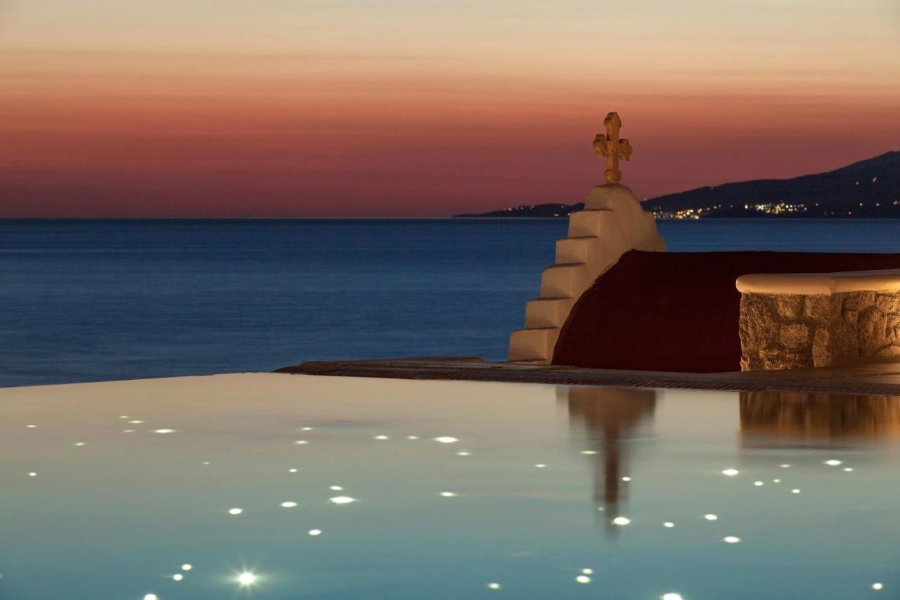 Bill & Coo Mykonos infinity pool at dusk with lit fiber optics reflected in water, white chapel with golden cross, and Aegean Sea at sunset