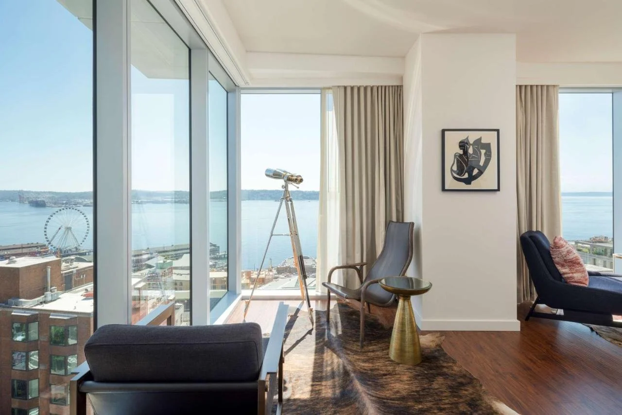 Thompson Seattle Seattle suite living area with floor-to-ceiling windows overlooking Elliott Bay, the Seattle Great Wheel, and Puget Sound, with a standing brass telescope in the foreground