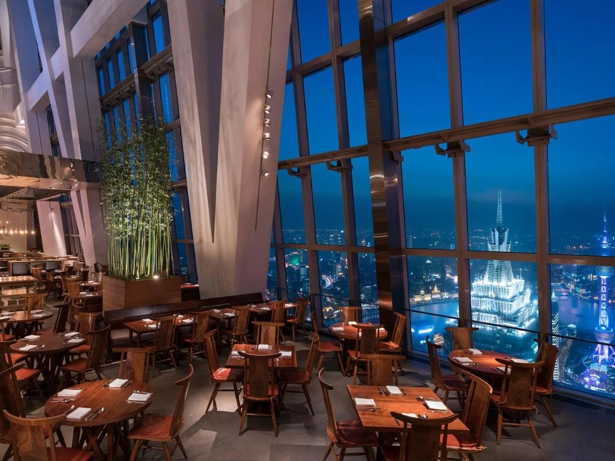Park Hyatt Shanghai restaurant at night with angled floor-to-ceiling windows overlooking the illuminated Bund waterfront and Huangpu River far below