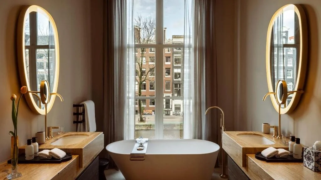 Rosewood Amsterdam bathroom with freestanding soaking tub between oval illuminated mirrors and window view of Prinsengracht canal houses