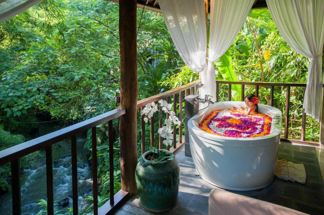 Komaneka at Bisma Ubud Bali open-air balcony bathtub filled with colorful flower petals overlooking rushing jungle river below