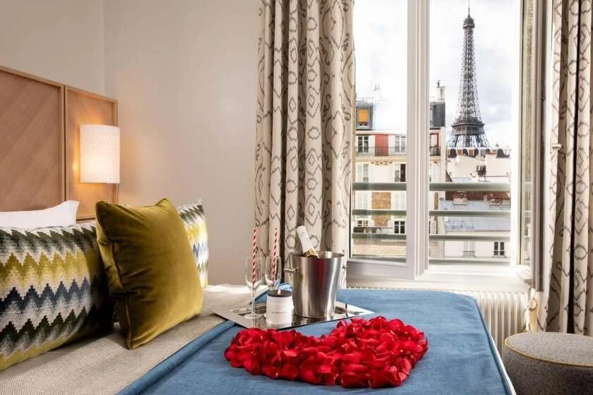 Les Jardins d’Eiffel Paris hotel room with champagne bucket and heart of rose petals on the bed, the Eiffel Tower framed in the window above the 7th arrondissement rooftops