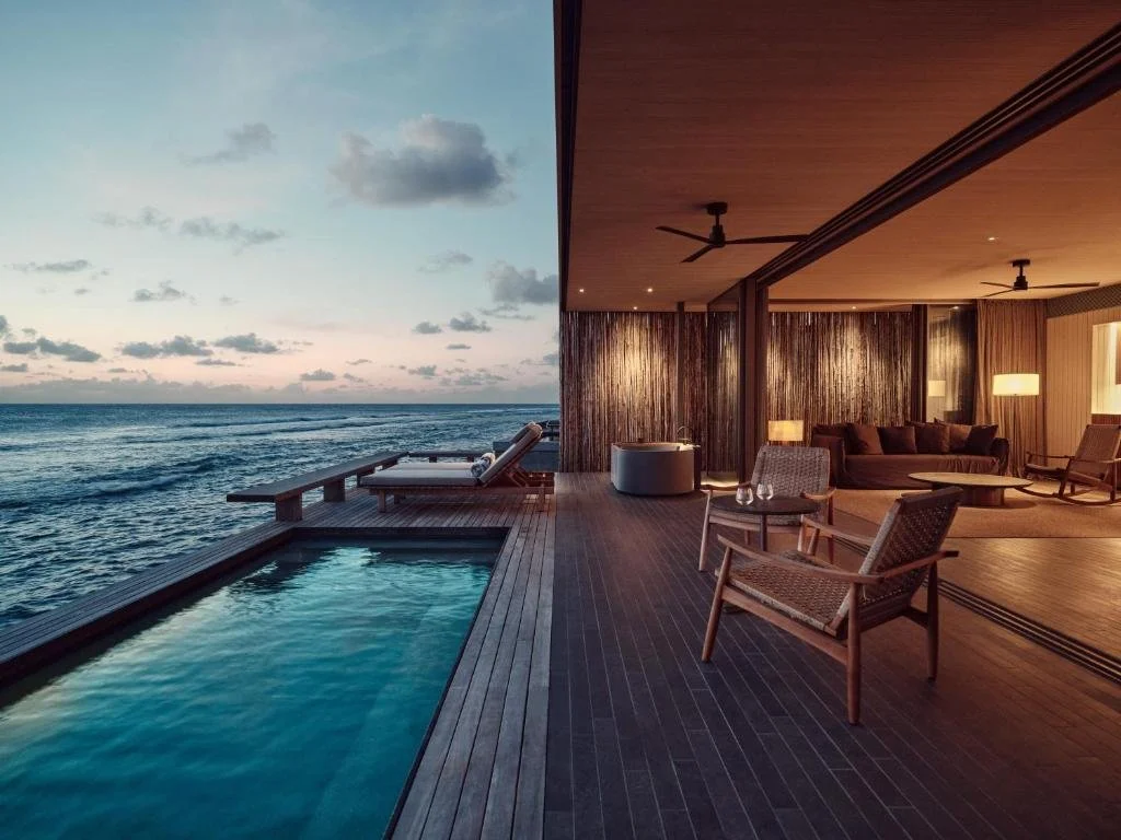 Patina Maldives Fari Islands overwater villa at dusk with private pool, open-plan living room with floor-to-ceiling glass walls facing ocean horizon at sunset