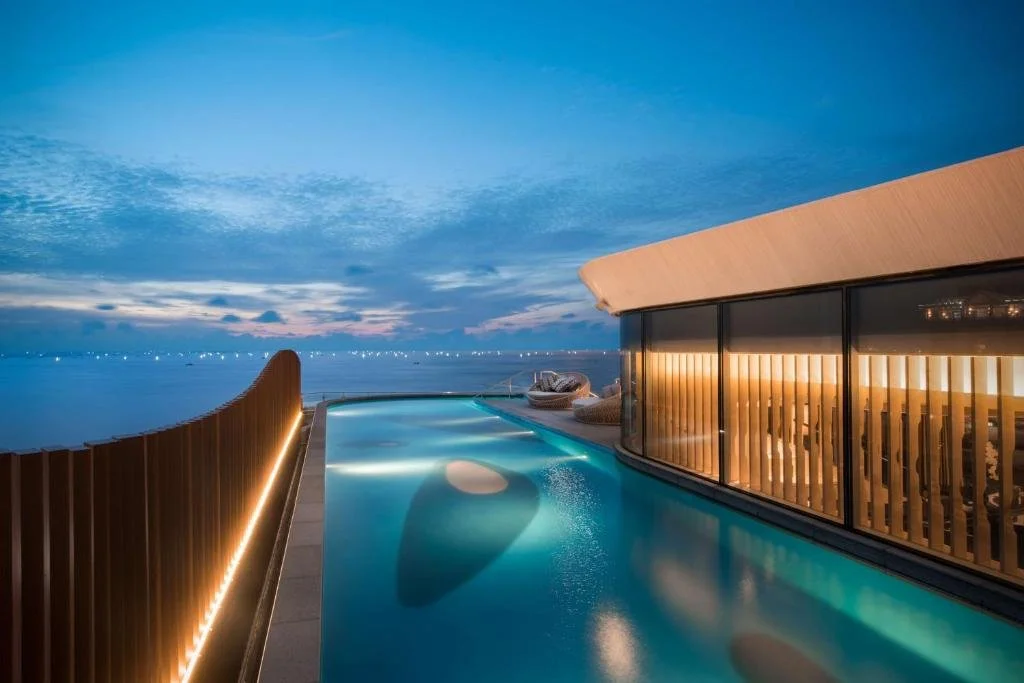 Conrad Manila Manila illuminated rooftop infinity pool at blue hour with Manila Bay stretching to the horizon and curved wooden façade reflected in the water
