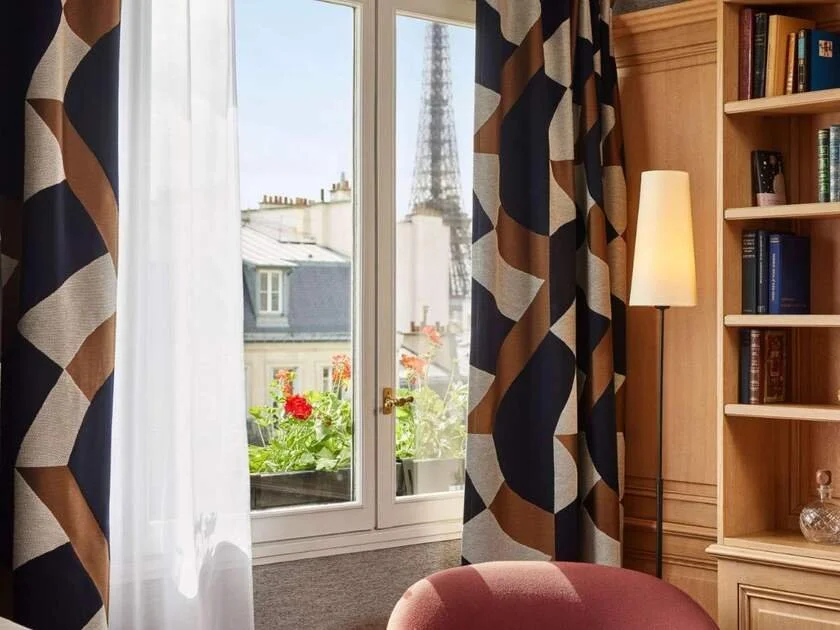 Sofitel Paris Baltimore Tour Eiffel hotel sitting area with geometric curtains, a wood bookcase, and red geraniums on the balcony, the Eiffel Tower partially visible above the 16th arrondissement rooftops