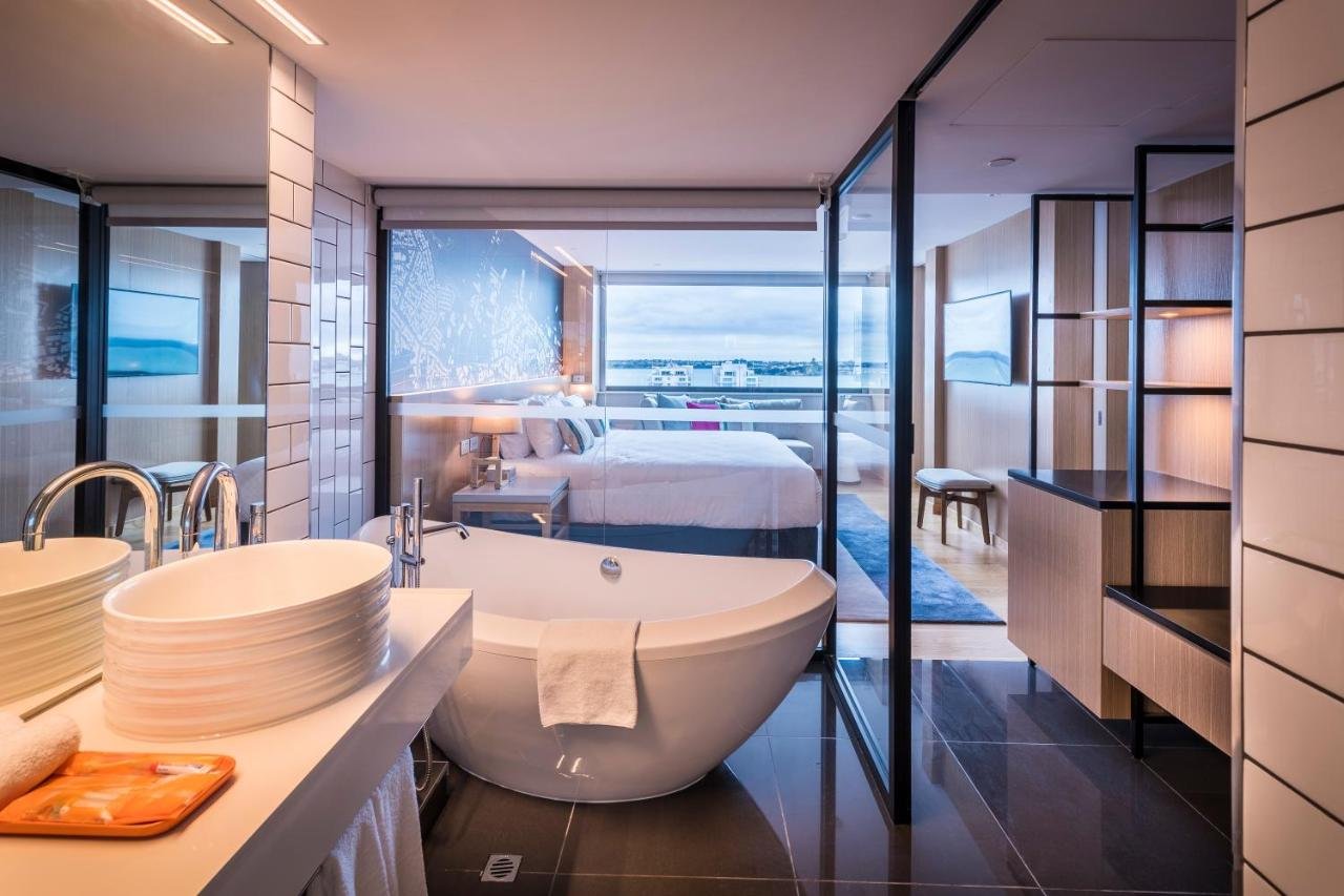 M Social Hotel Auckland Auckland suite with oval freestanding bathtub in the foreground and glass partition revealing bedroom windows facing the harbour at dusk