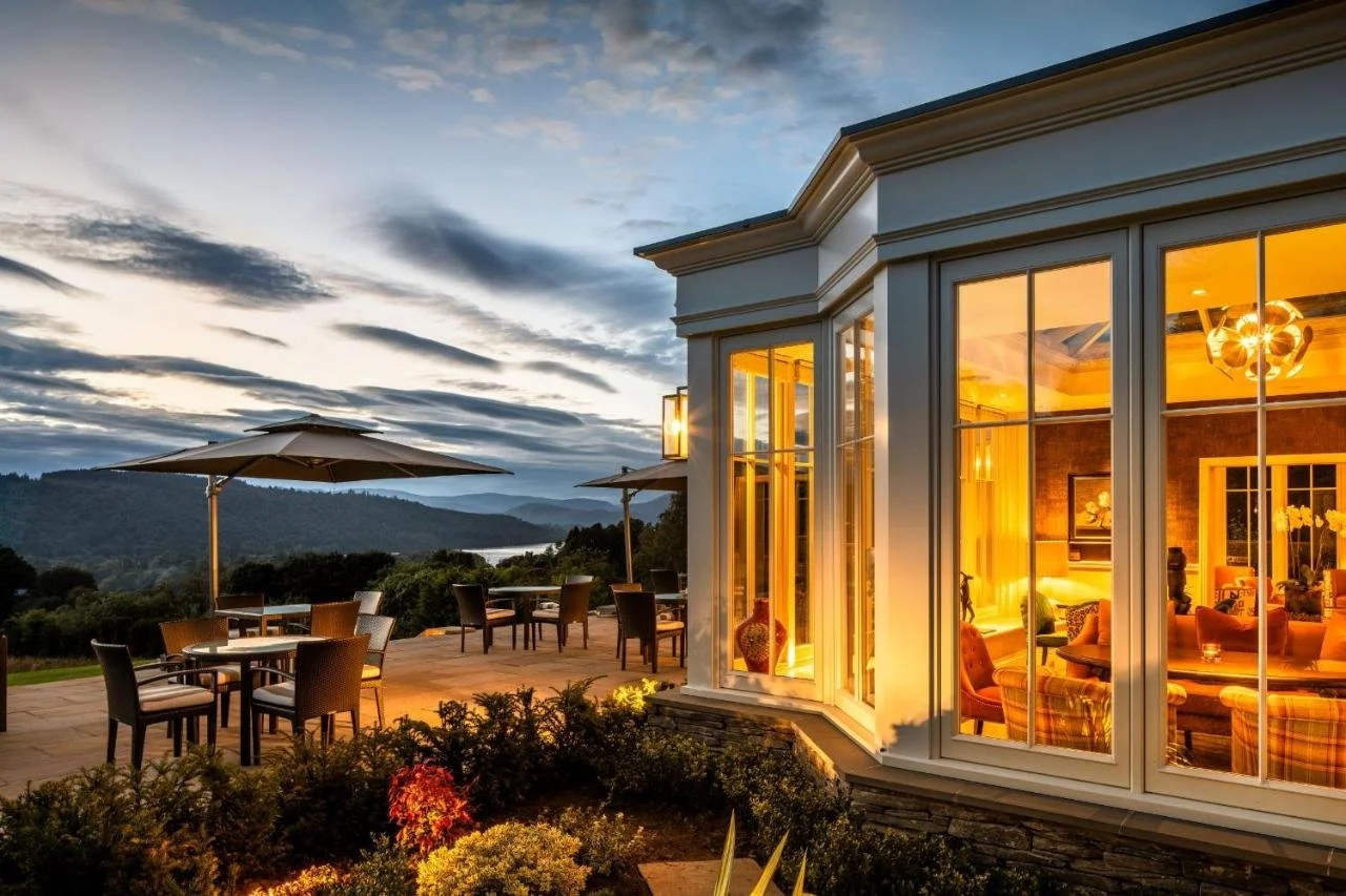 Linthwaite House Hotel Windermere hotel exterior and dining terrace at dusk with illuminated floor-to-ceiling windows and panoramic view over Lake Windermere and wooded hills