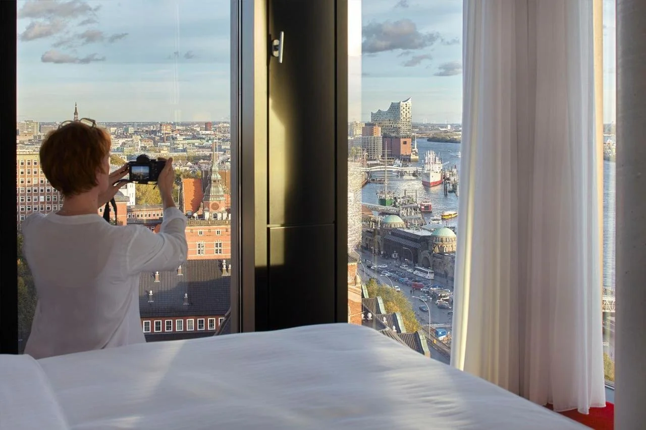 Empire Riverside Hotel Hamburg high-floor room with a guest photographing the view of the Elbe River, Elbphilharmonie, Landungsbrücken pier domes and container port through floor-to-ceiling windows