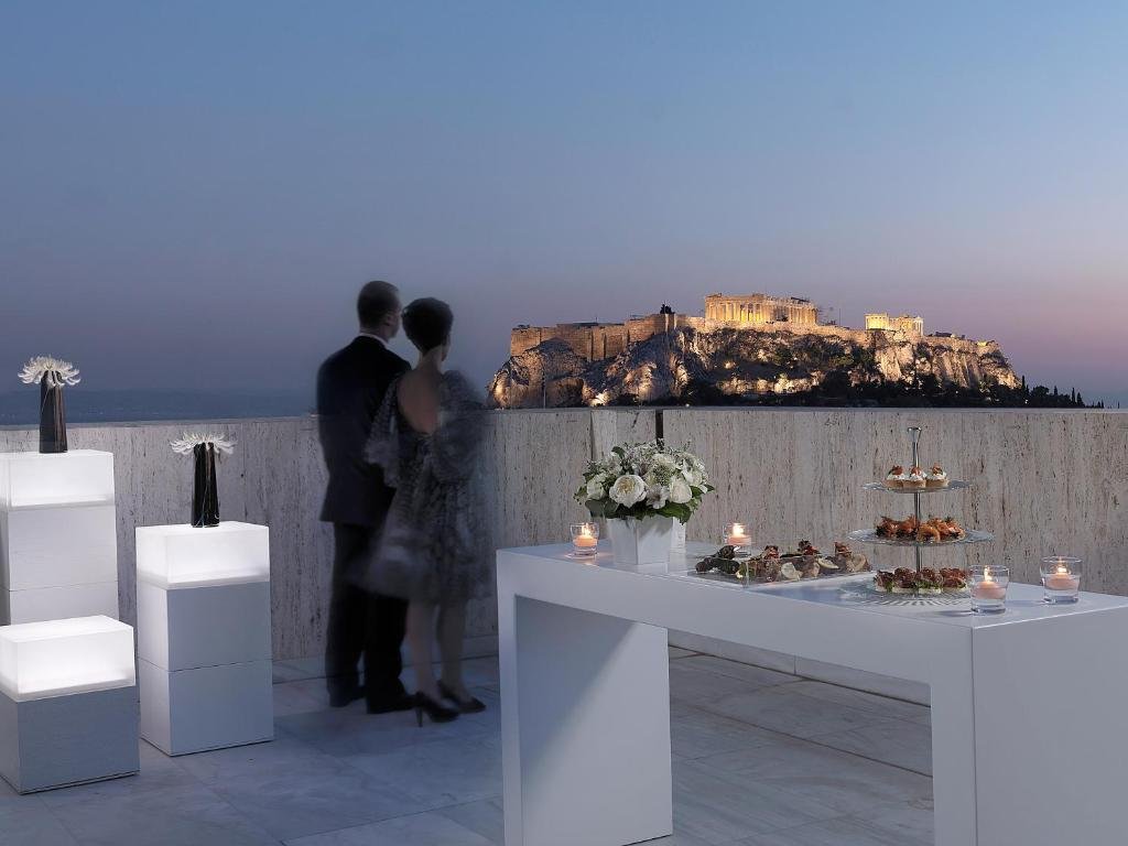 NJV Athens Plaza Athens rooftop terrace at dusk with couple at parapet overlooking illuminated Acropolis and white modern furniture
