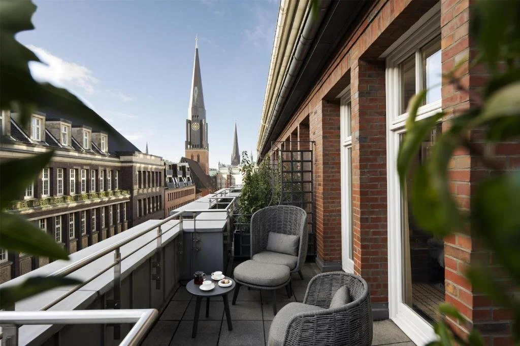 Conrad Hamburg hotel suite balcony with wicker seating overlooking Hamburg Altstadt rooftops, with the clock tower of Hauptkirche St. Jacobi rising prominently above the surrounding brick buildings