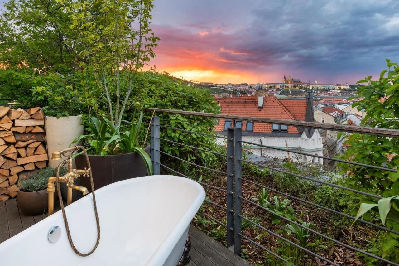 Mosaic House Design Hotel Prague outdoor freestanding bathtub on a rooftop terrace surrounded by greenery, Prague Castle visible against a stormy sunset sky