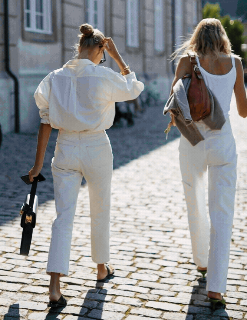 Two women walking on a cobblestone street in sunlight, carrying bags and one holding a smartphone.