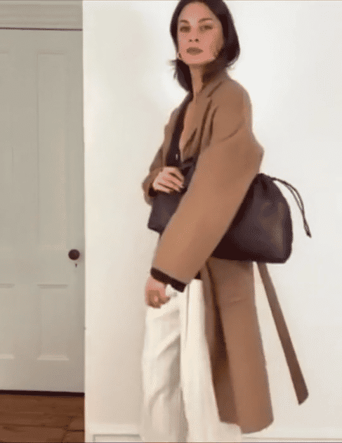 Woman carrying a black bag, wearing a tan coat, black top, and white pants standing indoors near a door.