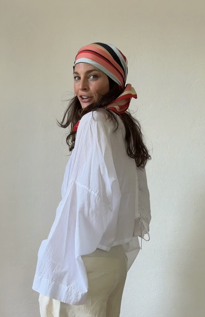 Woman wearing a colorful headscarf and white outfit, standing against a plain light-colored wall.