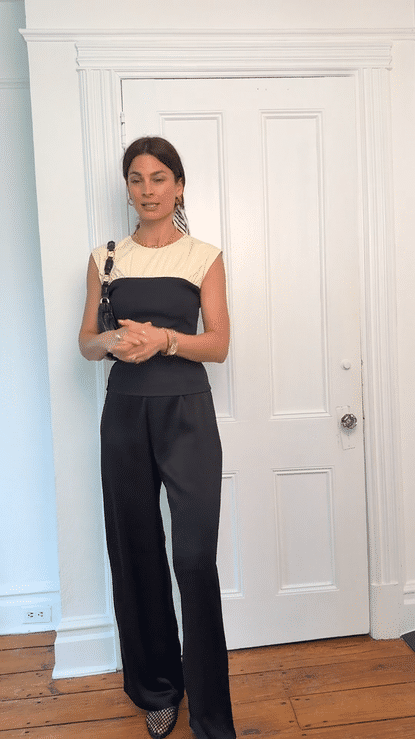 A woman stands indoors in front of a white door, wearing a black and beige sleeveless jumpsuit, with a handbag over her shoulder and jewelry.